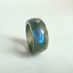 Puede incluir: Un anillo de labradorita facetada. El anillo tiene una banda ancha con un diseño geométrico. La piedra muestra un destello de color azul y verde. El anillo está sobre un fondo blanco.