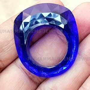 May include: A faceted, square-shaped, deep blue gemstone ring. The ring has a large, open centre and is held in a hand. The ring's surface reflects light, showcasing its cut. The text "MUHAMMAD GEMS JEWELS" is visible.