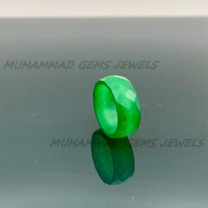 Könnte beinhalten: Ein facettierter, leuchtend grüner Ring. Der Ring ist ein massives Band mit polierter Innenseite und einer geometrischen, vielseitig facettierten Außenseite. Der Ring wird auf einer reflektierenden Oberfläche präsentiert. Der Text "MUHAMMAD GEMS JEWELS" ist sichtbar.