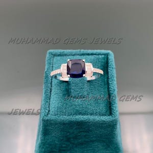 Può includere: Un anello d'argento con una grande pietra preziosa quadrata blu scuro al centro, affiancata da pietre rettangolari e trasparenti. L'anello è esposto su una superficie di velluto verde acqua. Il testo "MUHAMMAD GEMS JEWELS" è visibile.