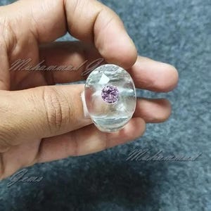 Può includere: Un anello ovale trasparente con una pietra preziosa viola incastonata al centro. L'anello è su una mano.