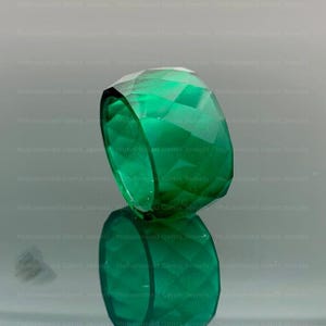 May include: A faceted, emerald-green ring. The gemstone ring is displayed on a reflective surface, showcasing its geometric cuts and vibrant color. The ring's design is bold and eye-catching.