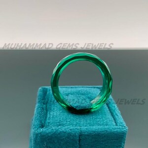 May include: A faceted, emerald green ring. The ring is displayed on a teal velvet surface. The text "MUHAMMAD GEMS JEWELS" is visible in the background.