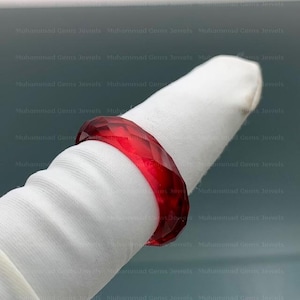 May include: A faceted, ruby-red ring. The ring is displayed on a white surface. The ring's design features multiple facets, creating a sparkling effect. The ring is a piece of jewelry.