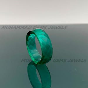May include: A faceted, emerald-green ring. The ring is made of a translucent material and reflects light, creating a sparkling effect. The ring is displayed on a reflective surface. The text "MUHAMMAD GEMS JEWELS" is visible.
