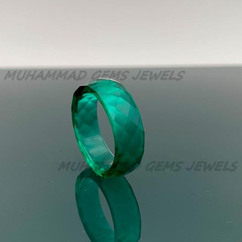 May include: A faceted, emerald-green ring. The ring is made of a translucent material and reflects light, creating a sparkling effect. The ring is displayed on a reflective surface. The text "MUHAMMAD GEMS JEWELS" is visible.