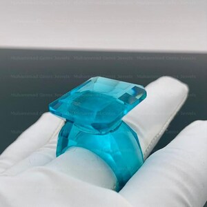 May include: A large, transparent, aquamarine-colored ring with a rectangular top and a rounded band. The ring is held by a gloved hand, showcasing its faceted design and vibrant color. The ring is a statement piece.