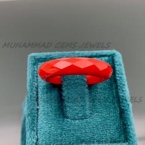 Könnte beinhalten: Ein facettierter, leuchtend roter Ring wird auf einer türkisfarbenen Samtoberfläche präsentiert. Der Ring hat ein geometrisches Design und ist der Mittelpunkt des Bildes. Der Text "MUHAMMAD GEMS JEWELS" ist im Hintergrund sichtbar.