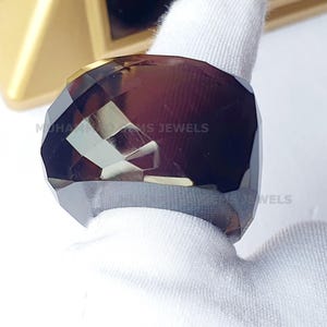 Puede incluir: Un anillo de piedra preciosa facetada de color morado oscuro con una banda dorada. El anillo está sobre un paño blanco.