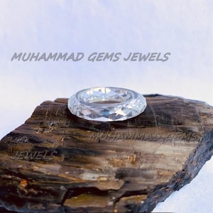 Peut inclure: Une bague transparente et facettée avec un motif géométrique. La bague est présentée sur un morceau de bois foncé et texturé. Le texte "MUHAMMAD GEMS JEWELS" est visible.
