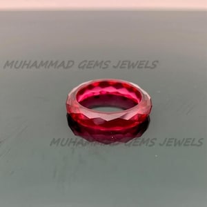 Peut inclure: Une bague en pierre précieuse rouge foncé à facettes. La bague présente un design géométrique avec de multiples facettes, créant un effet scintillant. La bague est posée sur une surface réfléchissante. Le texte "MUHAMMAD GEMS JEWELS" est visible.