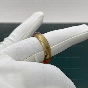 Op de afbeelding: Een goudkleurige ring met een gladde, afgeronde band. De ring wordt getoond op een witte gehandschoende hand. De ring lijkt een trouwring of een eenvoudig, elegant sieraad te zijn.