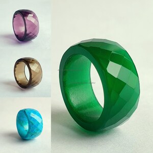 May include: A collection of gemstone rings in various colors. The rings include a faceted purple ring, a brown ring, a turquoise ring, and a large green ring. The rings are displayed on a white background.