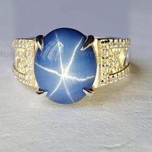 May include: A gold ring with a large, oval, blue star sapphire gemstone. The sapphire has a six-rayed star effect. The ring's band is decorated with smaller, clear gemstones, adding sparkle and detail to the jewellery.