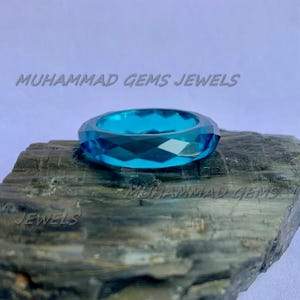 May include: A faceted, bright blue ring. The ring is set on a dark, textured surface. The text "MUHAMMAD GEMS JEWELS" is visible in the image.