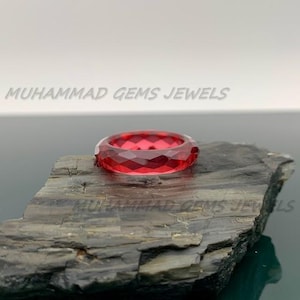 Puede incluir: Un anillo de piedra preciosa roja facetada. El anillo es de un color rojo rubí intenso y se encuentra sobre una superficie oscura y texturizada. El diseño del anillo presenta múltiples facetas que capturan la luz, creando un efecto brillante. El texto "MUHAMMAD GEMS JEWELS" está en el fondo.