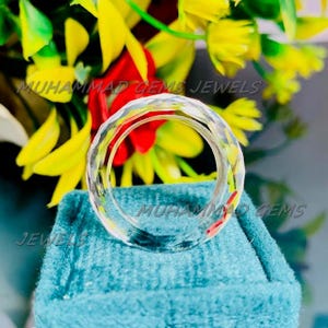 Può includere: Un anello trasparente e sfaccettato con un design circolare. L'anello è esposto su una superficie turchese, con uno sfondo di fiori gialli e rossi. Il testo "MUHAMMAD GEMS JEWELS" è visibile.
