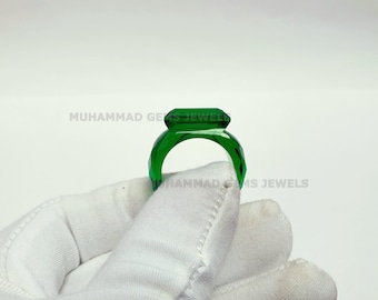 Puede incluir: Un anillo de vidrio verde con una piedra de forma rectangular. El anillo está sobre una superficie blanca.