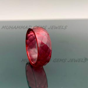 Könnte beinhalten: Ein facettierter, tiefroter Edelsteinring. Der Ring ist ein massives Band mit geometrischem Schliff, das Licht reflektiert. Der Ring wird auf einer reflektierenden Oberfläche präsentiert. Der Text "MUHAMMAD GEMS JEWELS" ist im Hintergrund sichtbar.