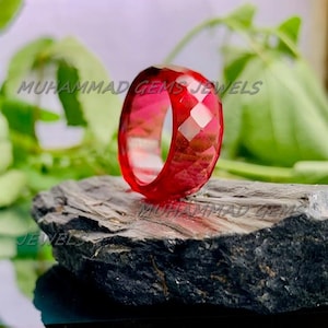 May include: A faceted, translucent red ring. The ring is displayed on a dark, textured surface with green foliage in the background. The text "MUHAMMAD GEMS JEWELS" is visible.