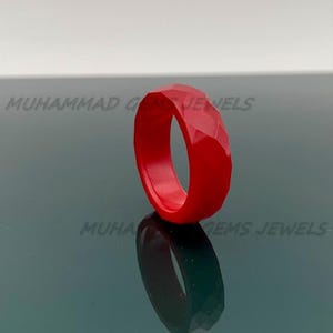 Könnte beinhalten: Ein leuchtend roter Ring mit facettiertem Design. Die Oberfläche des Rings ist in mehrere flache Ebenen unterteilt, wodurch ein geometrisches Muster entsteht. Der Ring wird auf einer reflektierenden Oberfläche mit seiner Reflexion darunter gezeigt. Ein Schmuckstück.