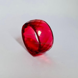 May include: A faceted, deep red gemstone ring. The ring is a vibrant, transparent red with multiple facets that catch the light. The ring is set against a white background, highlighting its colour and shape. The ring appears to be a statement piece.