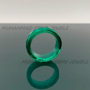 May include: A faceted, emerald-green ring. The ring is made of a transparent material and has a geometric design. The ring is displayed on a reflective surface. The text "MUHAMMAD GEMS JEWELS" is visible in the background.