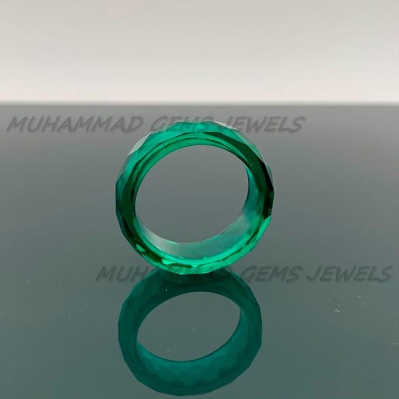 May include: A faceted, emerald-green ring. The ring is made of a transparent material and has a geometric design. The ring is displayed on a reflective surface. The text "MUHAMMAD GEMS JEWELS" is visible in the background.