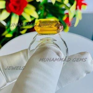 Könnte beinhalten: Ein klarer Ring mit einem großen, rechteckigen, gelben Edelstein. Der Ring wird von einer behandschuhten Hand gehalten. Der Text "MUHAMMAD GEMS JEWELS" ist sichtbar.