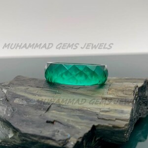 May include: A faceted, emerald-green ring. The ring is displayed on a dark, textured surface. The words "MUHAMMAD GEMS JEWELS" are printed in white across the top of the image.