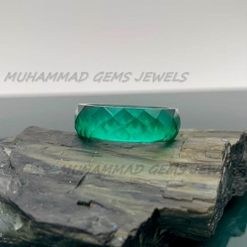 May include: A faceted, emerald-green ring. The ring is displayed on a dark, textured surface. The words "MUHAMMAD GEMS JEWELS" are printed in white across the top of the image.