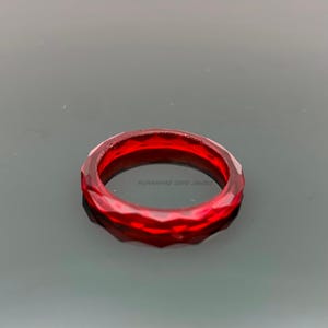 May include: A faceted, deep red ring. The ring has a geometric design with multiple facets, creating a sparkling effect. The ring is made of a translucent material, and the text "MUHAMMAD GEMS JEWELS" is visible.