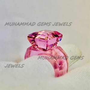 Könnte beinhalten: Ein pinkfarbener Edelsteinring mit einem großen, rautenförmigen Mittelstück. Der Ring liegt auf einer weißen Oberfläche, mit dem Text "MUHAMMAD GEMS JEWELS" im Hintergrund.