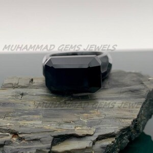 Könnte beinhalten: Ein schwarzer Edelsteinring mit einem großen, rechteckig geschliffenen Stein. Der Ring liegt auf einer dunklen, strukturierten Oberfläche. Der Text "MUHAMMAD GEMS JEWELS" ist im Hintergrund sichtbar.