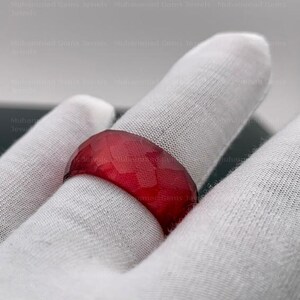 May include: A faceted, deep red gemstone ring. The ring's surface is cut with multiple facets, creating a textured appearance. The ring is set on a white surface.
