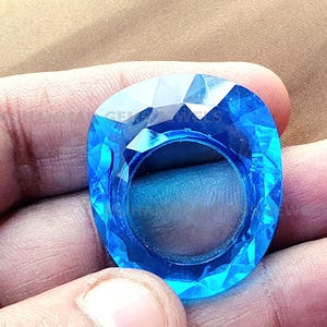 May include: A large, faceted, blue gemstone ring. The ring is made of clear glass and has a faceted design. The ring is held in a hand.