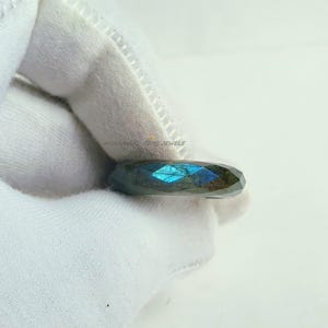 May include: A faceted labradorite ring with a dark grey band and iridescent blue and green flashes. The ring is held against a white background. The text "MUHAMMAD GEMS JEWELS" is visible.