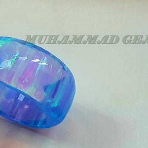 May include: A blue opal ring with iridescent hues of purple and green. The ring is held by tweezers against a white background. Text in the background reads "MUHAMMAD GEMS JEWELS".
