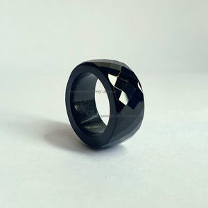 Könnte beinhalten: Ein facettierter schwarzer Ring mit glänzender Oberfläche. Der Ring hat ein breites Band und ein geometrisches Design. Er ist vor einem weißen Hintergrund platziert. Der Ring ist ein Schmuckstück.