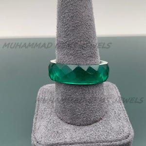 May include: A faceted, emerald-green ring is displayed on a gray ring holder. The ring has a geometric design with multiple facets, creating a sparkling effect. The ring is a bold statement piece.