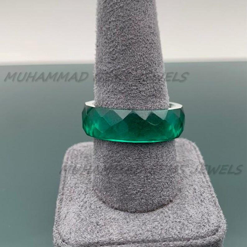 May include: A faceted, emerald-green ring is displayed on a gray ring holder. The ring has a geometric design with multiple facets, creating a sparkling effect. The ring is a bold statement piece.