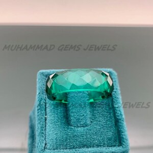 May include: A faceted, emerald-green gemstone ring. The ring is set on a teal velvet display. The text "MUHAMMAD GEMS JEWELS" is visible in the background. The gemstone has a rectangular shape with multiple facets.