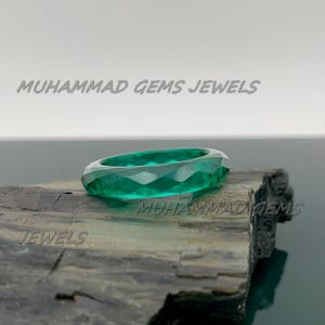 May include: A faceted emerald green ring. The ring is set on a dark, textured surface. The text "MUHAMMAD GEMS JEWELS" is visible in the background.