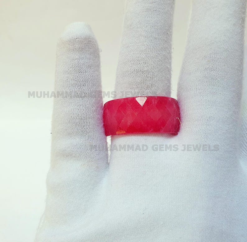 May include: A faceted, bright red ring is displayed on a white gloved hand. The ring has a geometric design and a flat top. The text "MUHAMMAD GEMS JEWELS" is visible on the glove.