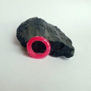 Pode incluir: Um anel rosa brilhante facetado com um design circular. O anel está colocado contra uma rocha cinza escura sobre um fundo branco. A superfície do anel reflete a luz, mostrando seus cortes geométricos. É uma joia.