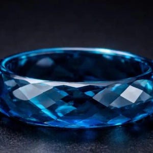 Puede incluir: Un anillo de piedra preciosa azul facetado. El anillo es de un tono azul vibrante, con múltiples facetas que capturan la luz. El anillo es de estilo banda, con un interior liso y pulido. El anillo está sobre un fondo oscuro.