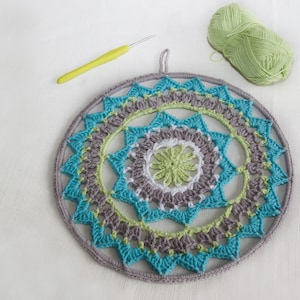 Könnte beinhalten: Ein gehäkelter Mandala-Wandbehang mit einem kreisförmigen Design in den Farben Türkis, Grün, Grau und Weiß. Das Mandala ist mit verschiedenen Maschen gearbeitet und hat ein zartes, filigranes Muster.