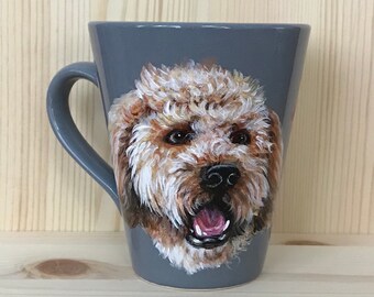 Puede incluir: Una taza de cerámica gris con un retrato pintado a mano de un perro marrón y blanco. El perro tiene un pelaje esponjoso y mira directamente al espectador.