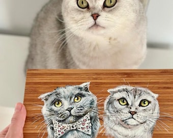 Op de afbeelding: Een houten snijplank met twee handgeschilderde portretten van grijze tabby-katten. Eén kat draagt een roze en witte strik met hartjes. De plank heeft de tekst "IKEA 23145 1720" onderaan gedrukt.