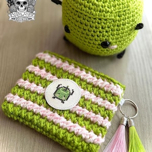 May include: A lime green crocheted apple plush with black eyes and a stem, alongside a rectangular crocheted pouch with alternating lime green and light pink stripes. The pouch features a circular patch with a green pixelated character and two tassels.